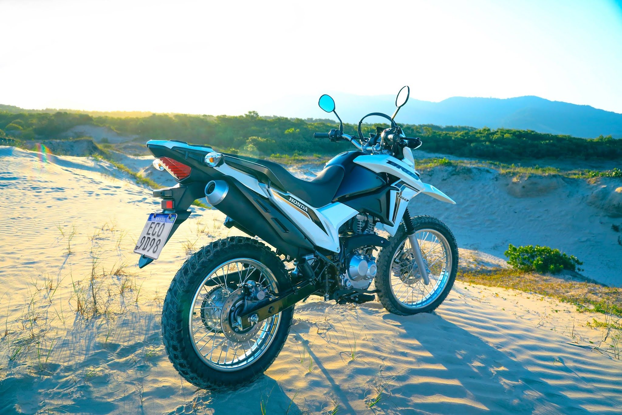 Visão completa da motocicleta Honda NXR 160 2023 na cor branca, estacionada na praia